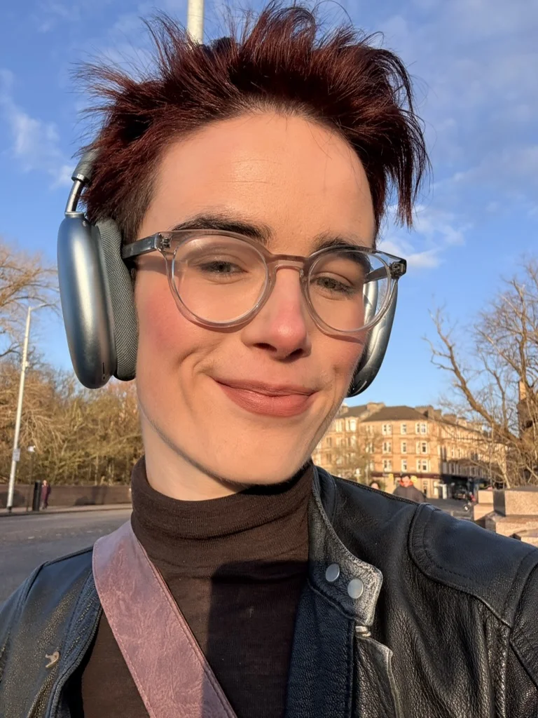 Lou outside in the sun in Glasgow, wearing headphones and clear glasses